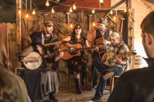 Small group of musicians playing guitar, banjo, bass, and washboard in front of rustic scene with an audience