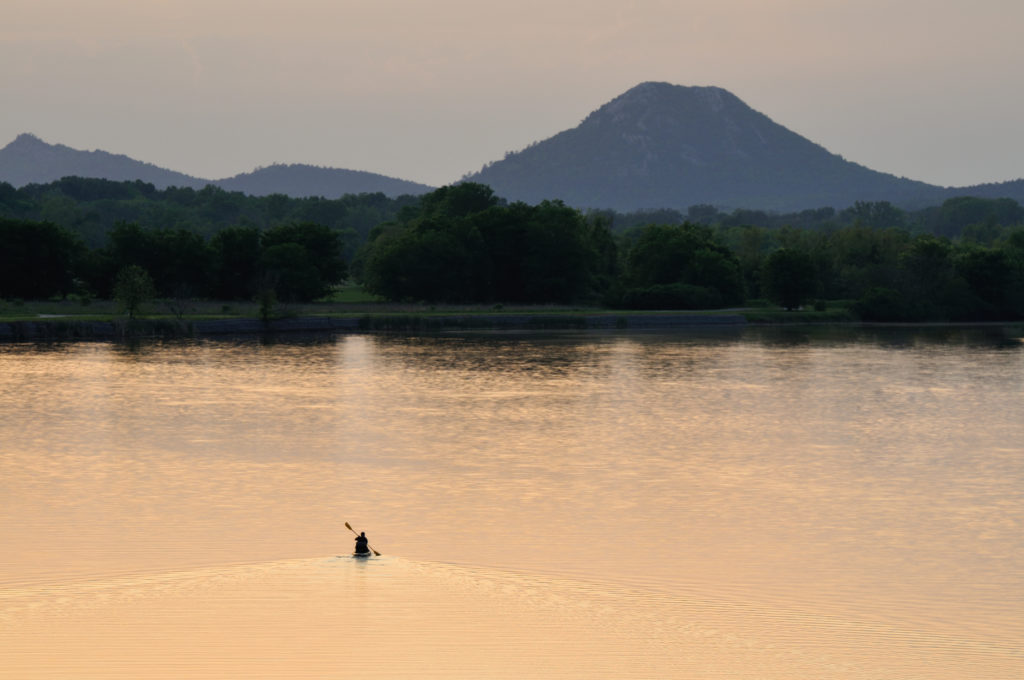 Lone person canoeing on a lake at sunset or sunrise with mountain in the background.